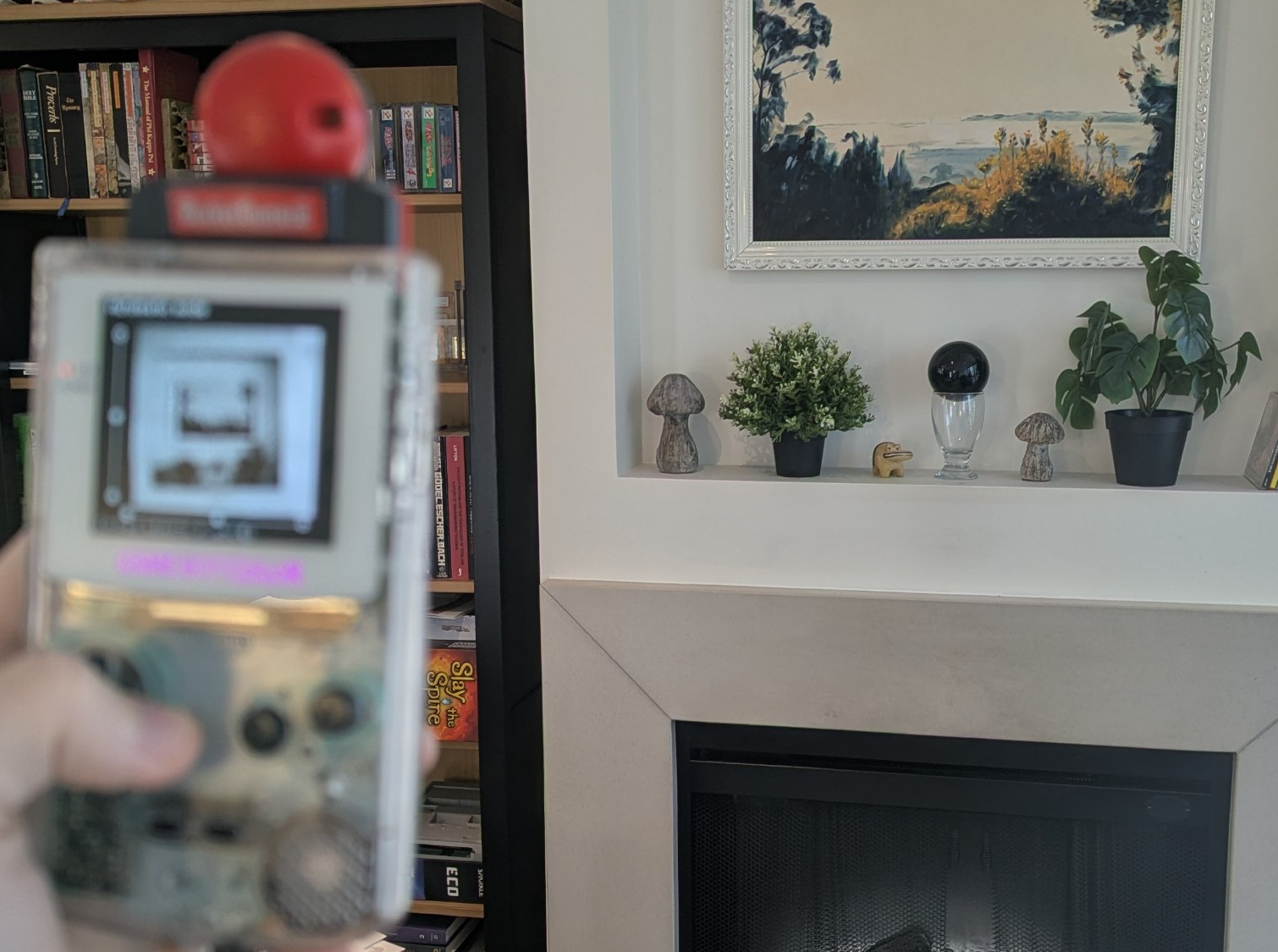 Hand holding a Game Boy Color with a PicNRec cartridge, framing a fireplace mantel. The GBC screen shows a dithered preview of the scene.